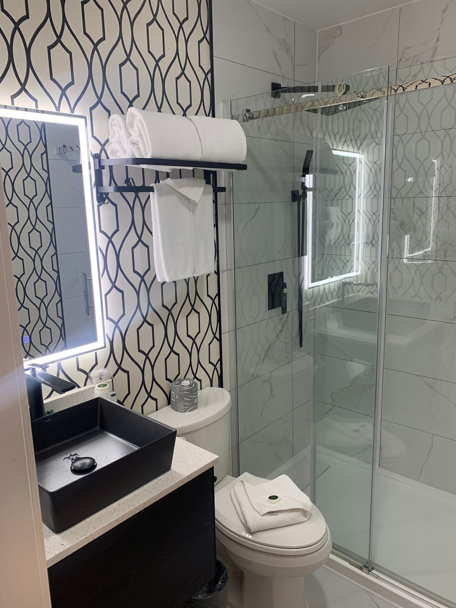 A modern bathroom with a geometric patterned wall, a glass shower enclosure, a white toilet, and a black sink with a mirror above it.