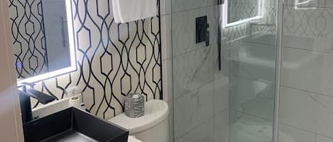 A modern bathroom with a geometric patterned wall, a glass shower enclosure, a white toilet, and a black sink with a mirror above it.