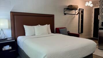 Un lit bien fait, avec une tête de lit marron, une table de chevet avec une lampe et une étagère fixée au mur où est posée une serviette.