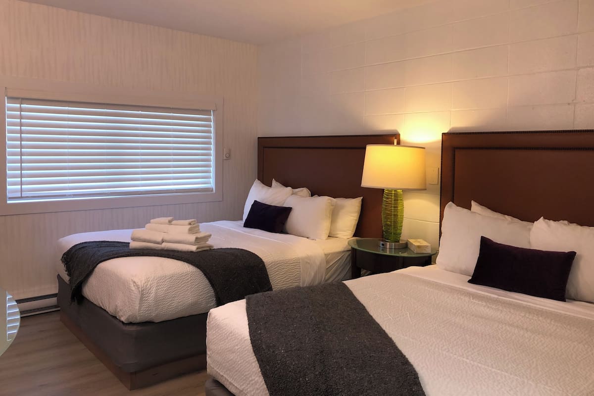 Una habitación de hotel con dos camas, una ventana con persianas, una lámpara de noche y una mesita.