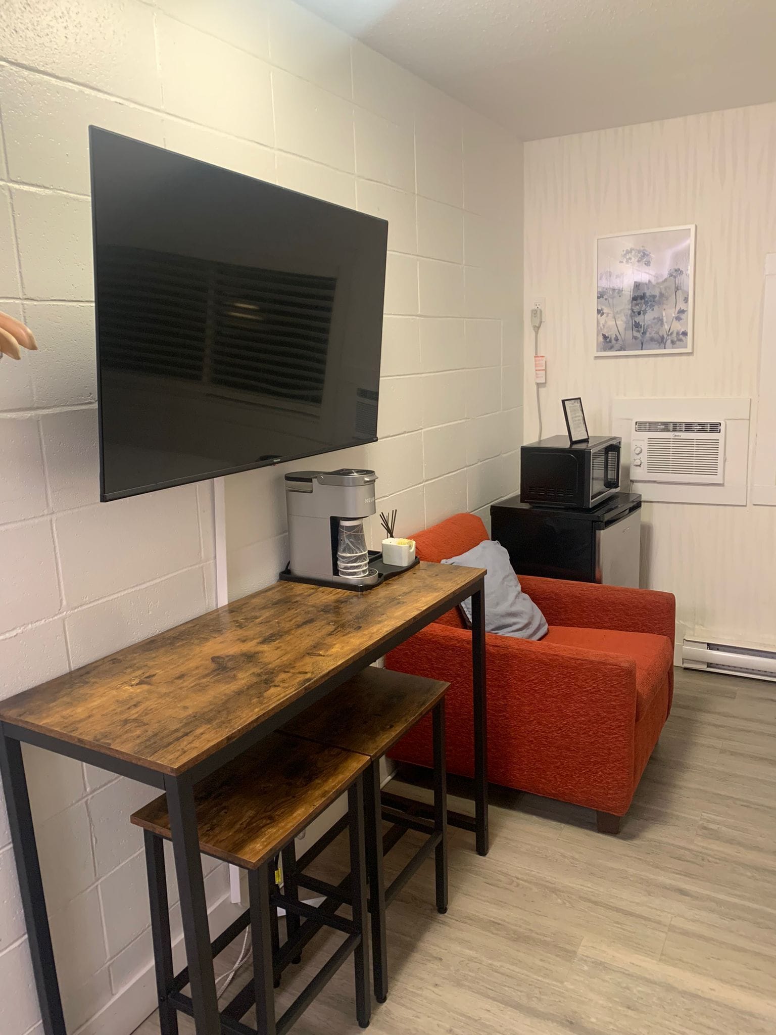 A compact living space with a wooden table and bench, a flat-screen TV mounted on the wall, a small kitchenette with a microwave, and a red sofa.