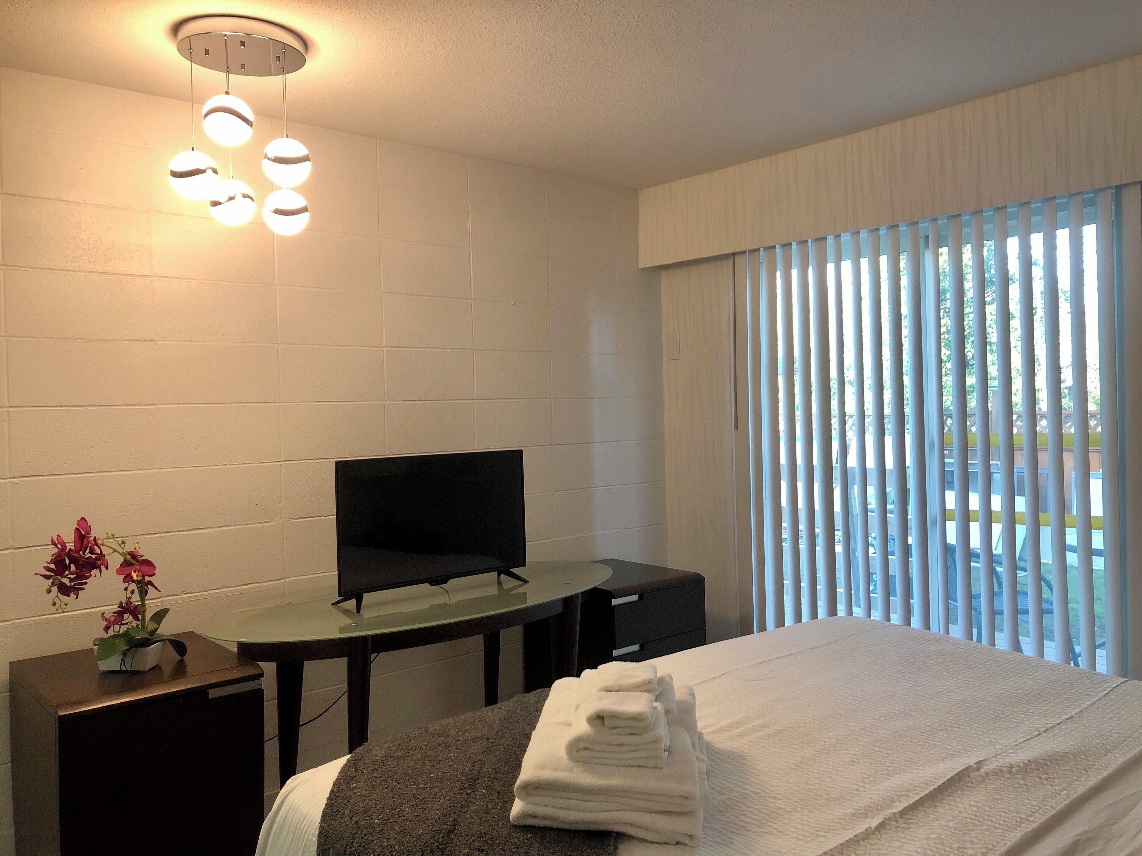 A hotel room with a bed, a TV on a stand, a small table, and a window with blinds.
