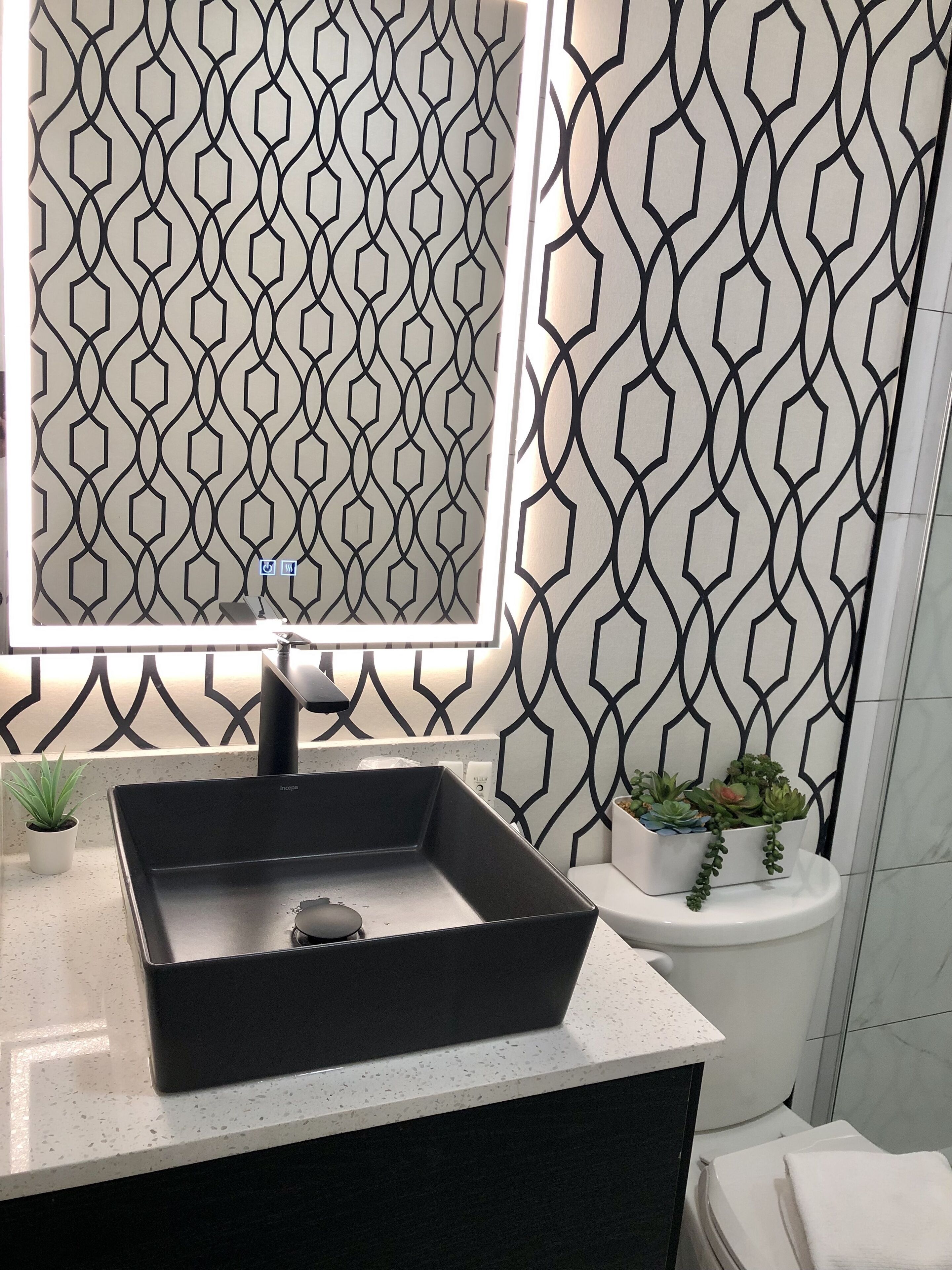 A bathroom with a black sink, a mirror with integrated lighting, a potted plant, and a toilet.