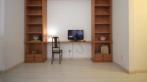 Quarto com televisão sobre suporte de madeira, uma cadeira e prateleiras com diversos objetos.