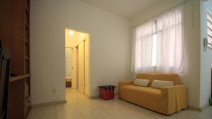 Quarto com guarda-roupa de madeira, sofá amarelo, lixeira vermelha e janela com cortinas brancas.