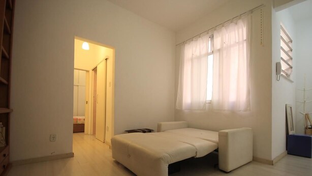 Um quarto pequeno e luminoso com uma cama, uma janela com cortinas leves e uma porta que dá para outro cômodo.