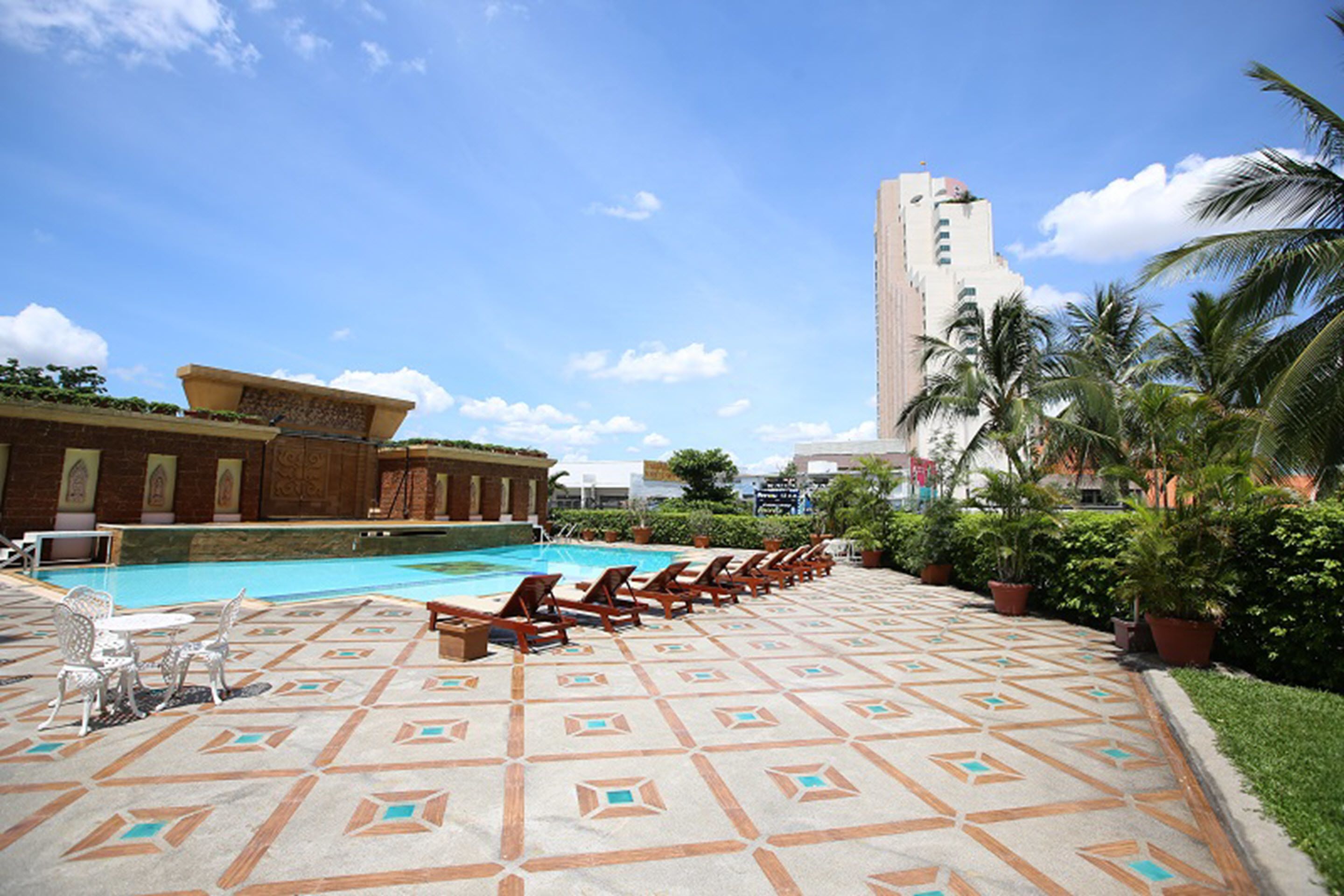 A pool area with lounge chairs and a patterned floor, surrounded by greenery and a tall building in the background.