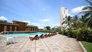 A pool area with lounge chairs and a patterned floor, surrounded by greenery and a tall building in the background.