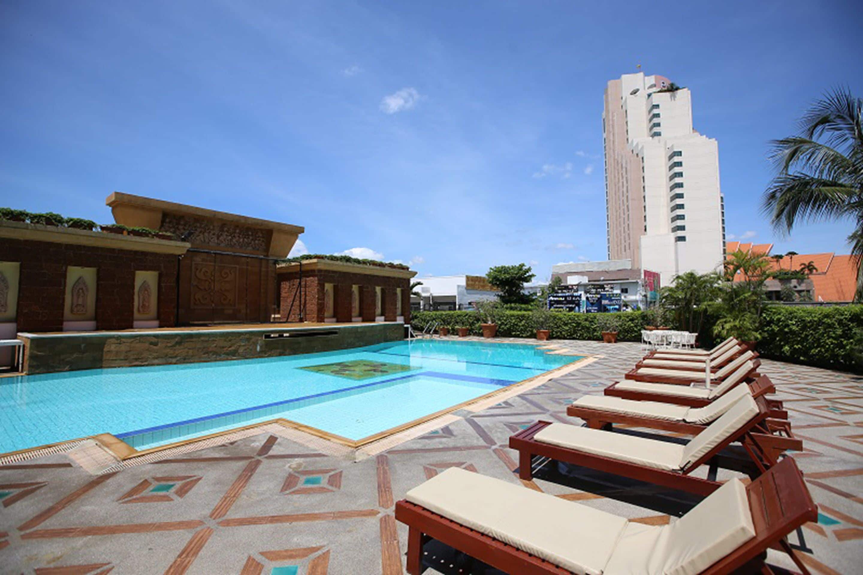 Una piscina en la azotea con sillones de descanso, piso estampado y un edificio alto al fondo.