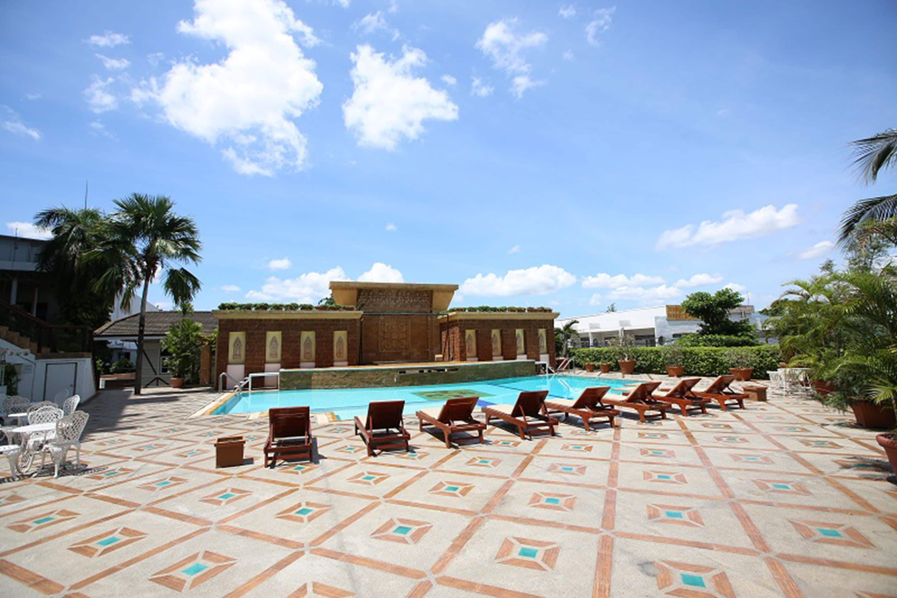 A pool area with lounge chairs and a patterned floor, surrounded by palm trees and a building with a tiled facade.