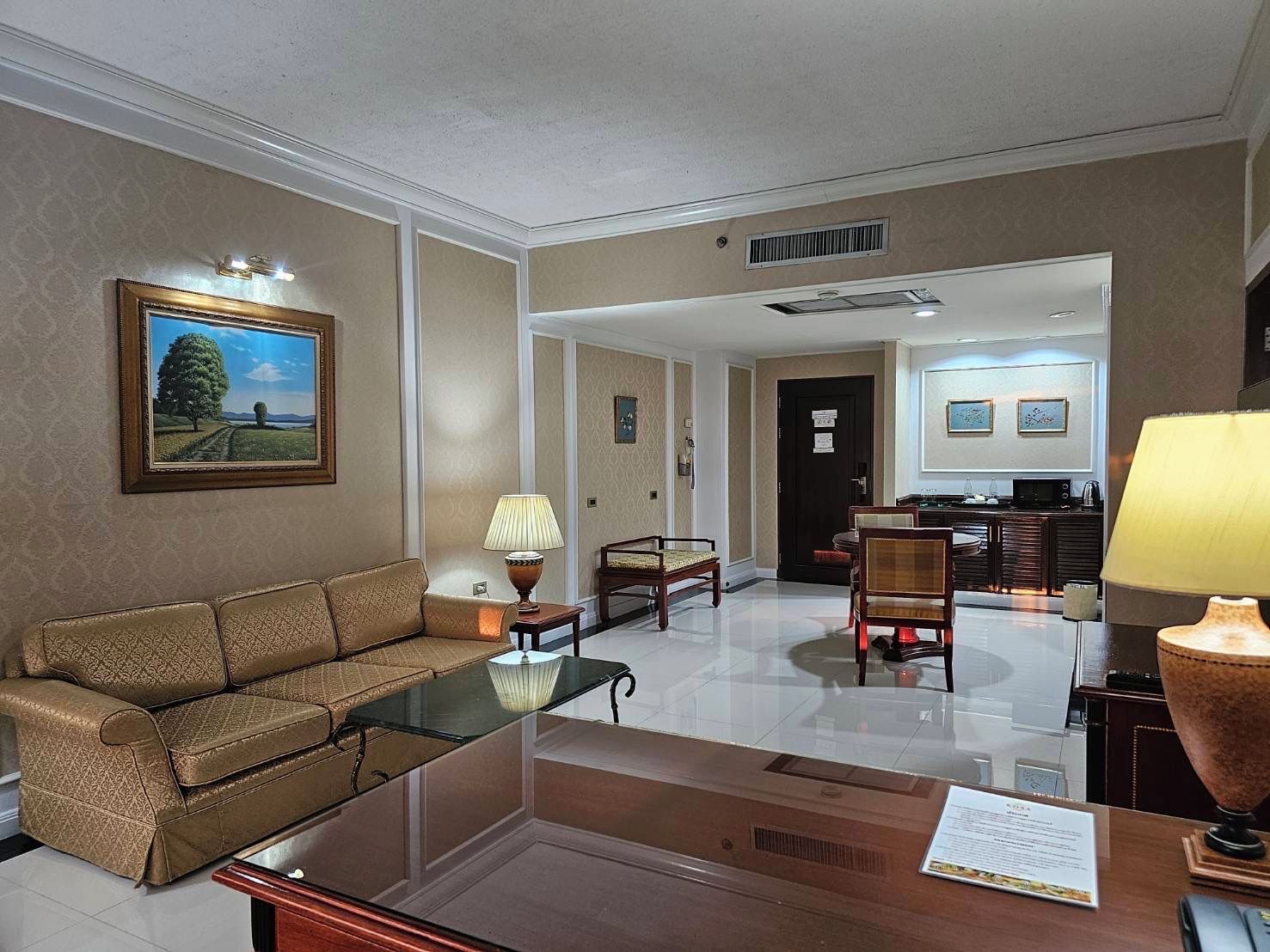 Habitación de hotel con sofá, una mesita de vidrio, lámpara, mesa de comedor con sillas, zona de cocina y un cuadro en la pared.