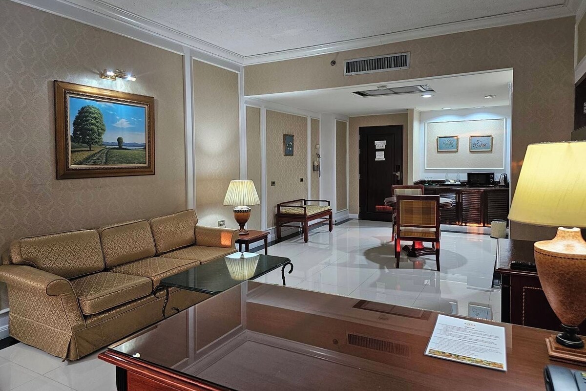 A hotel room with a sofa, a glass coffee table, a lamp, a desk with a chair, and a kitchen area in the background.