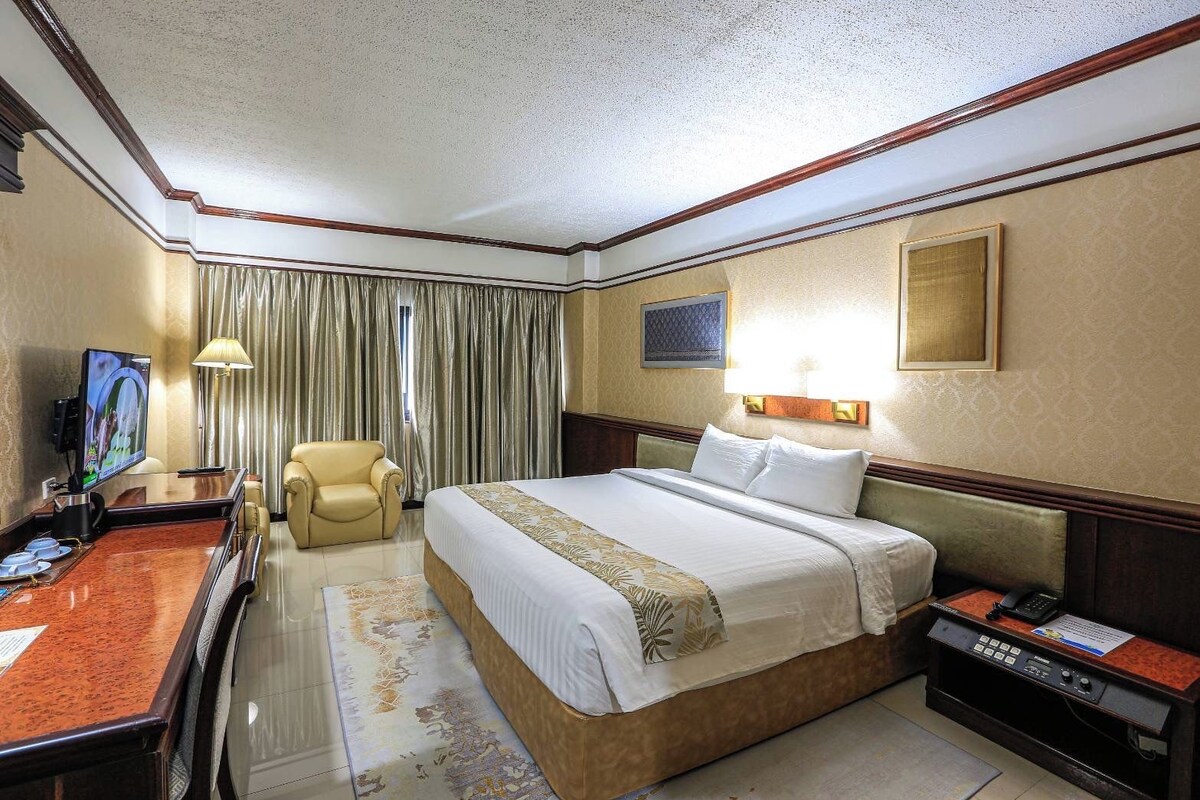 A hotel room with a bed, a desk with a television, a chair, a desk lamp, and a bedside table with a phone and a sign.