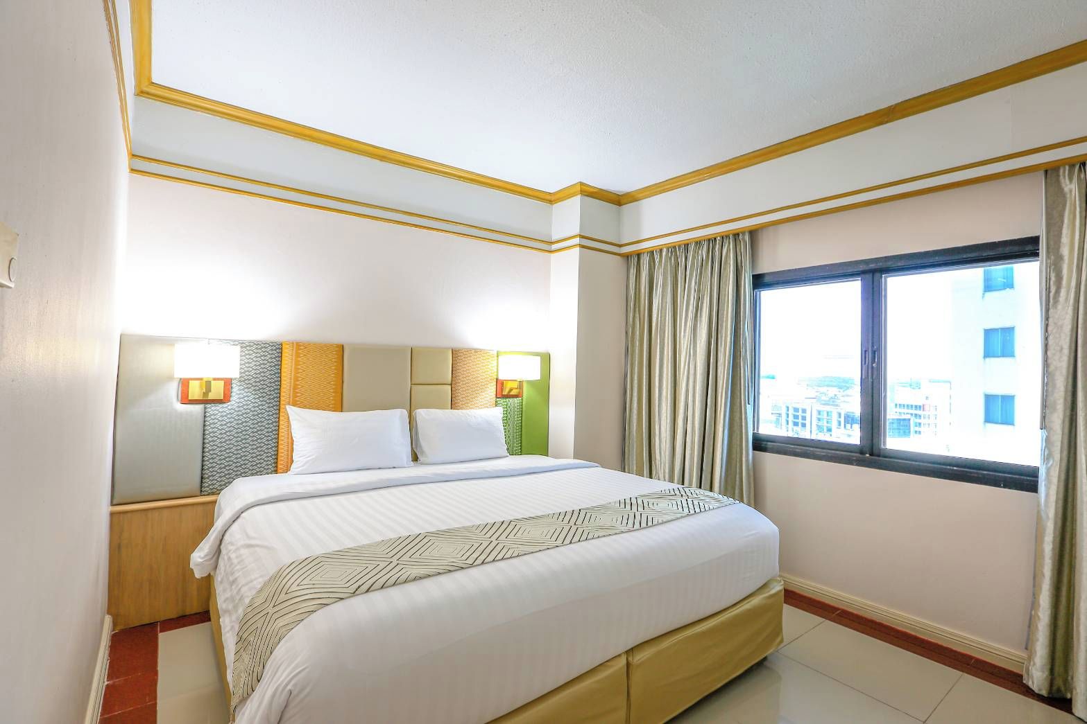 A hotel room with a bed, a nightstand, a lamp, a window with curtains, and a view of buildings outside.