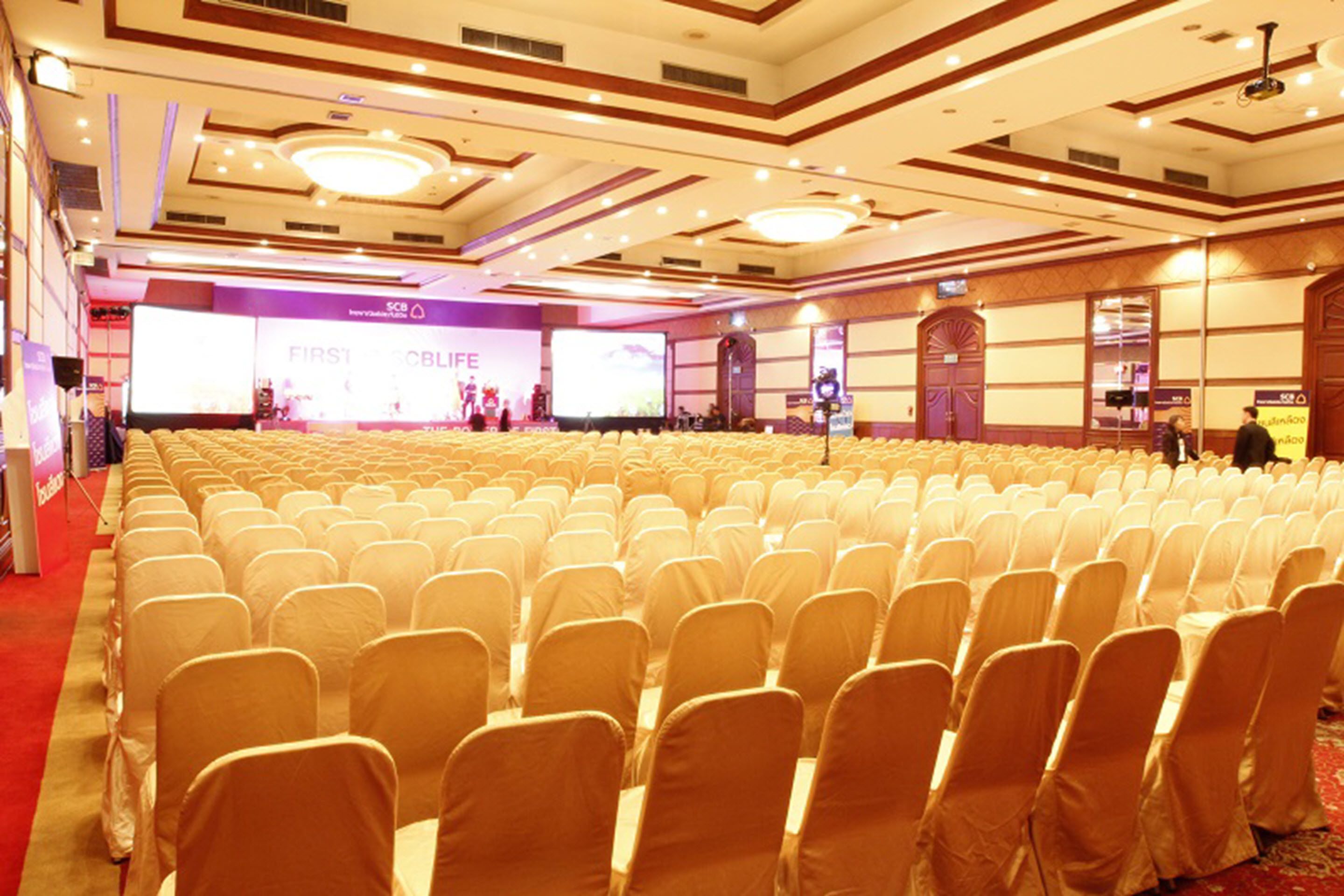 A conference room with rows of chairs covered in white chair covers, a stage with a large screen, and a banner reading "FIRST CBLE".