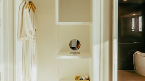 A minimalist bathroom with a white vanity, a hanging robe, and a shelf with toiletries. - Mantra Varee Hotel (Khon Kaen)