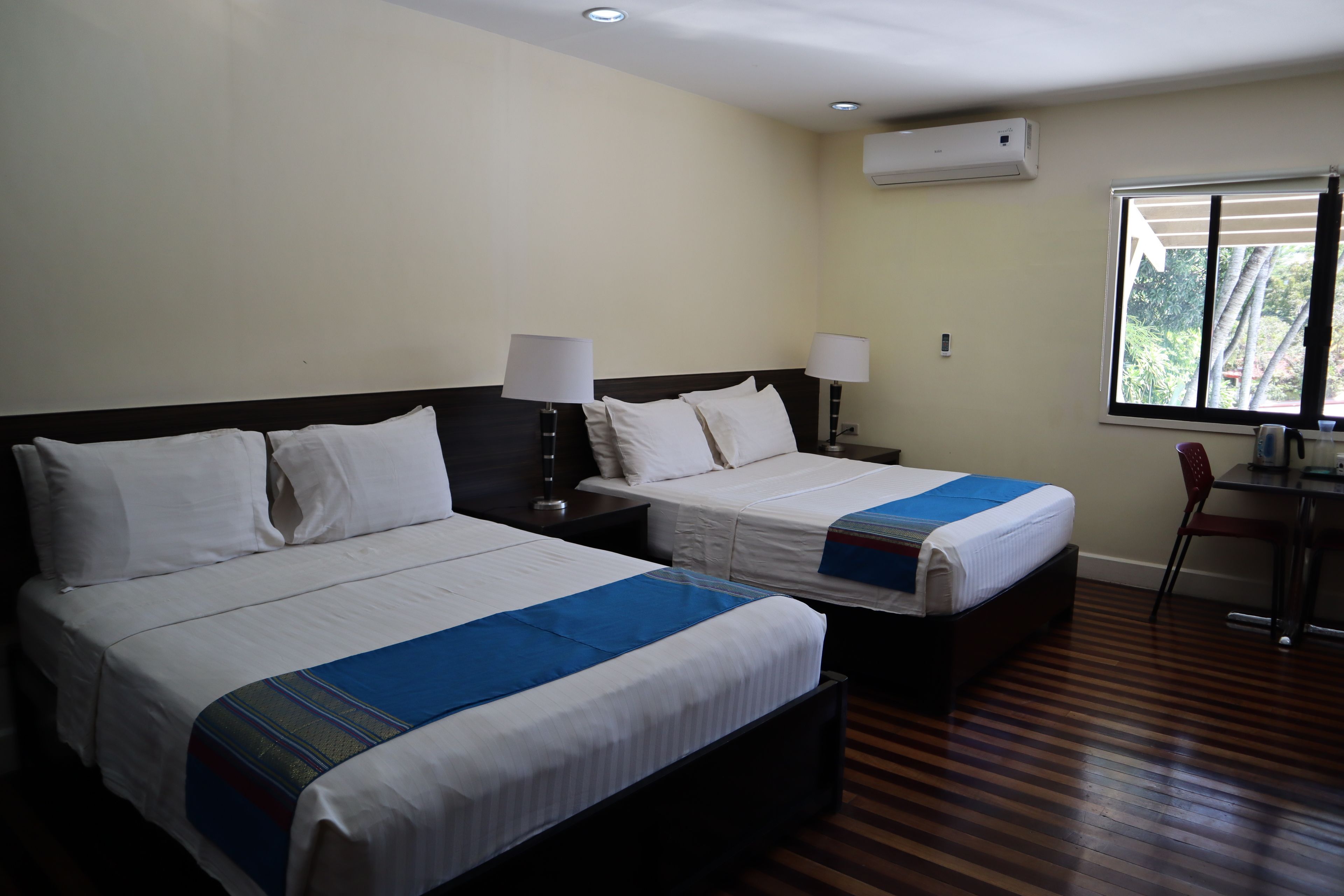 A hotel room with two beds, each with a white bedspread and blue accents, a desk with a chair, and a window with a view of greenery.