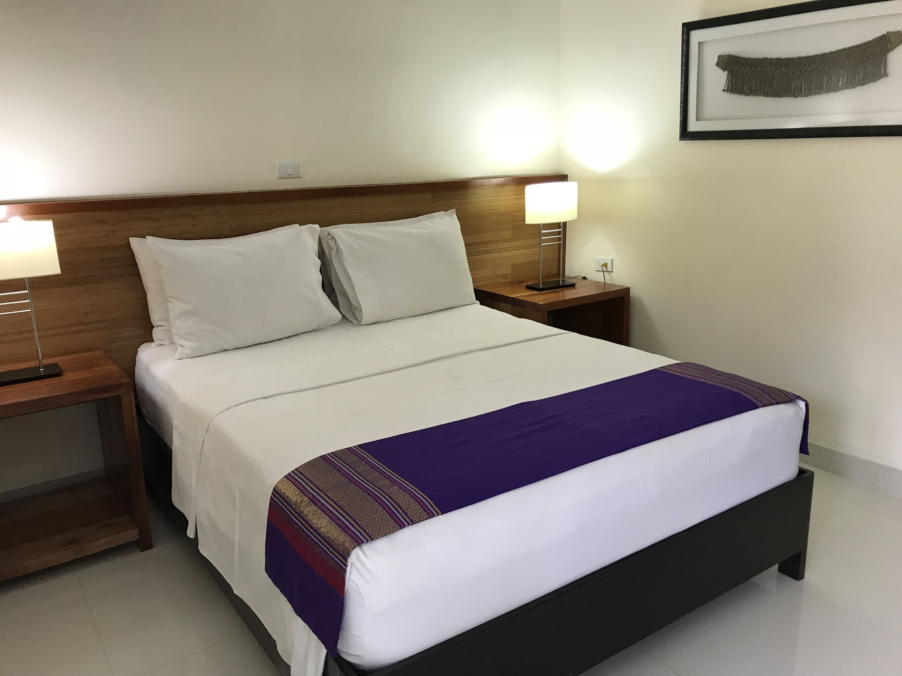A neatly made bed with white linens and a purple and gold striped blanket. There are two bedside tables with lamps, and a framed picture on the wall.