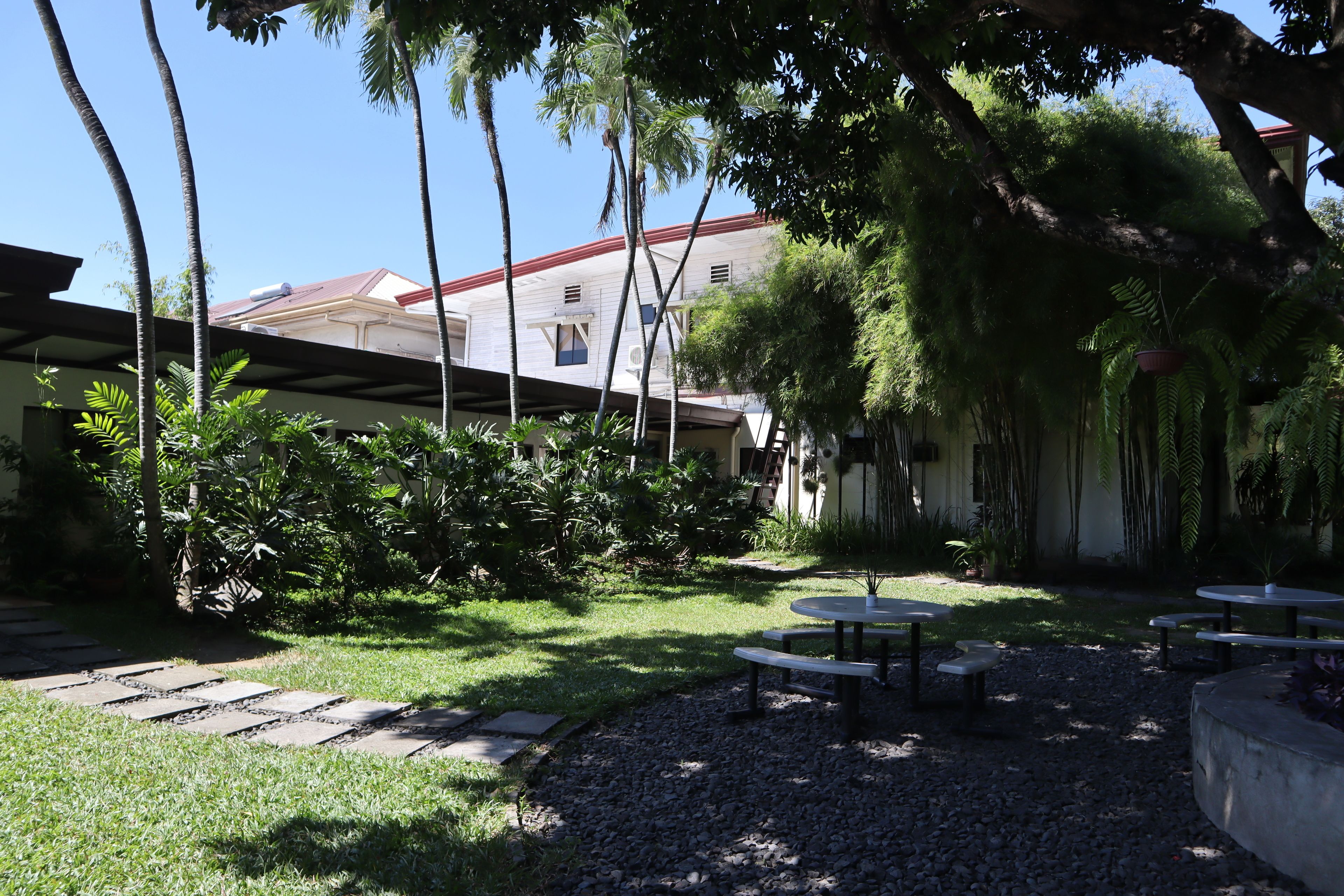 A courtyard with a covered seating area, palm trees, and a building with a red roof in the background.