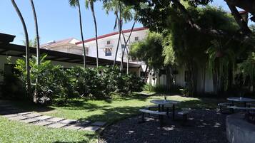 A courtyard with a covered seating area, palm trees, and a building with a red roof in the background.