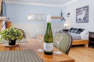 Quarto com mesa de jantar posta com taças de vinho e uma garrafa. Há uma planta em vaso sobre a mesa, uma cama com manta e um quadro na parede.