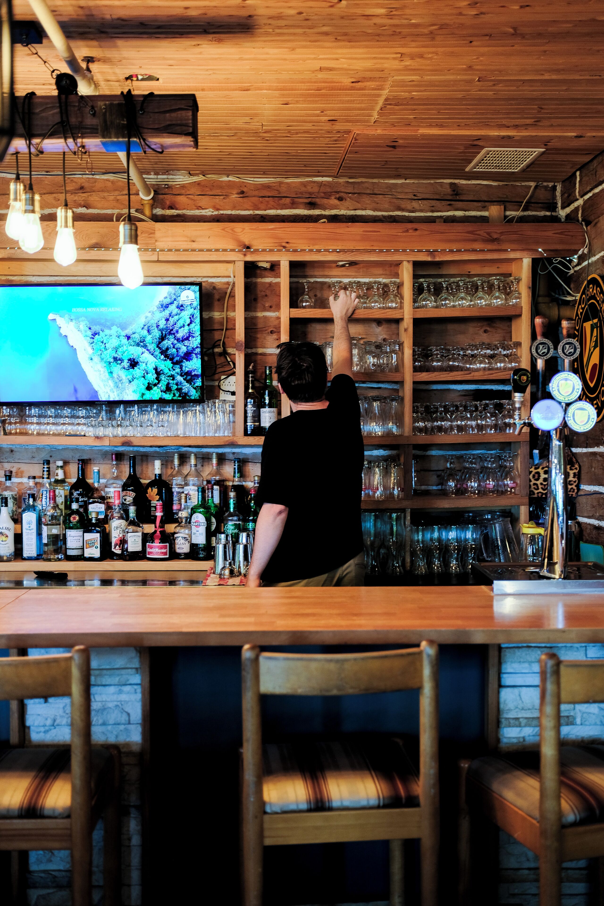 A bar area with a bartender, shelves of liquor bottles, and a television displaying a nature scene.