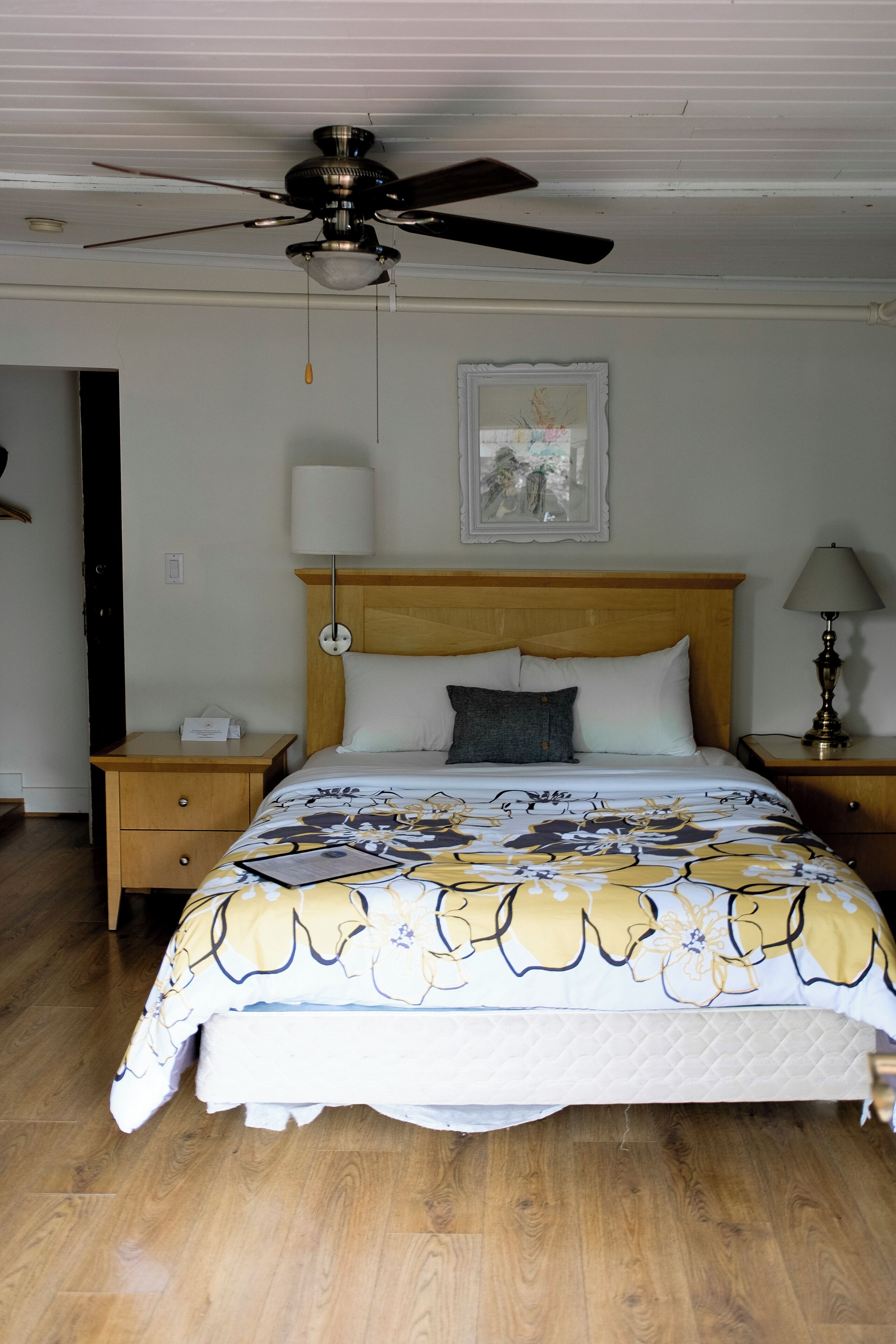 Un dormitorio con una cama de madera, un ventilador de techo, una mesita de noche, una lámpara de noche y un cuadro en la pared.