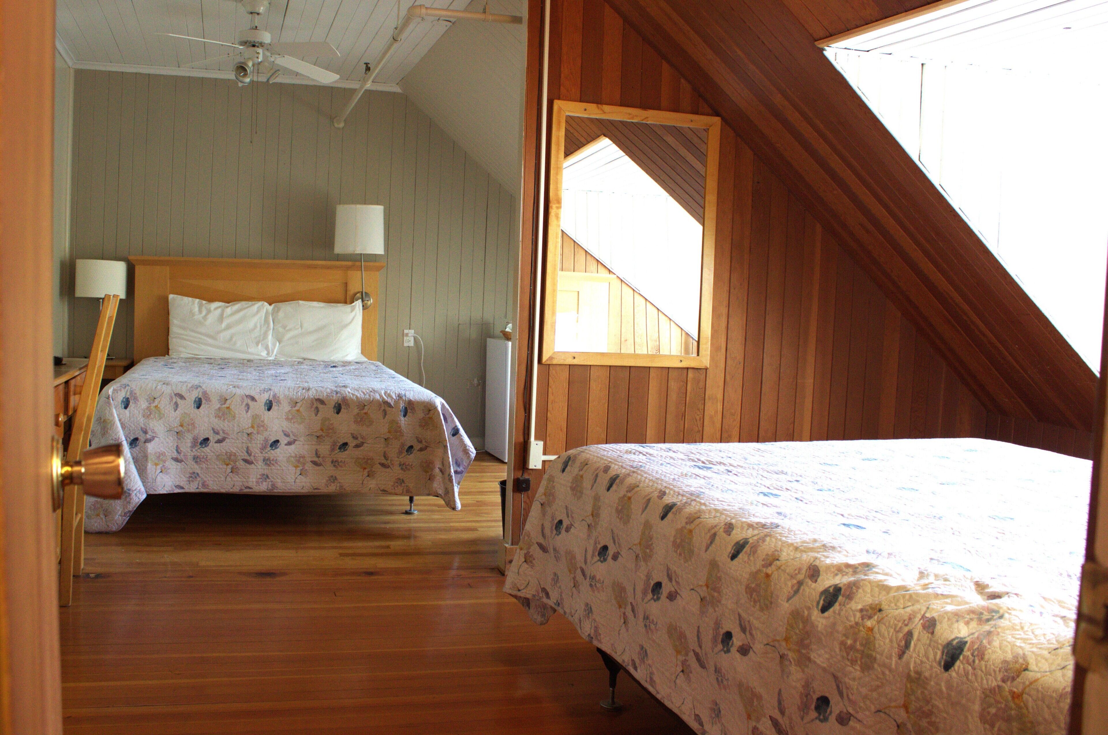 A bedroom with two beds, wooden flooring, a ceiling fan, and a window with a wooden frame.