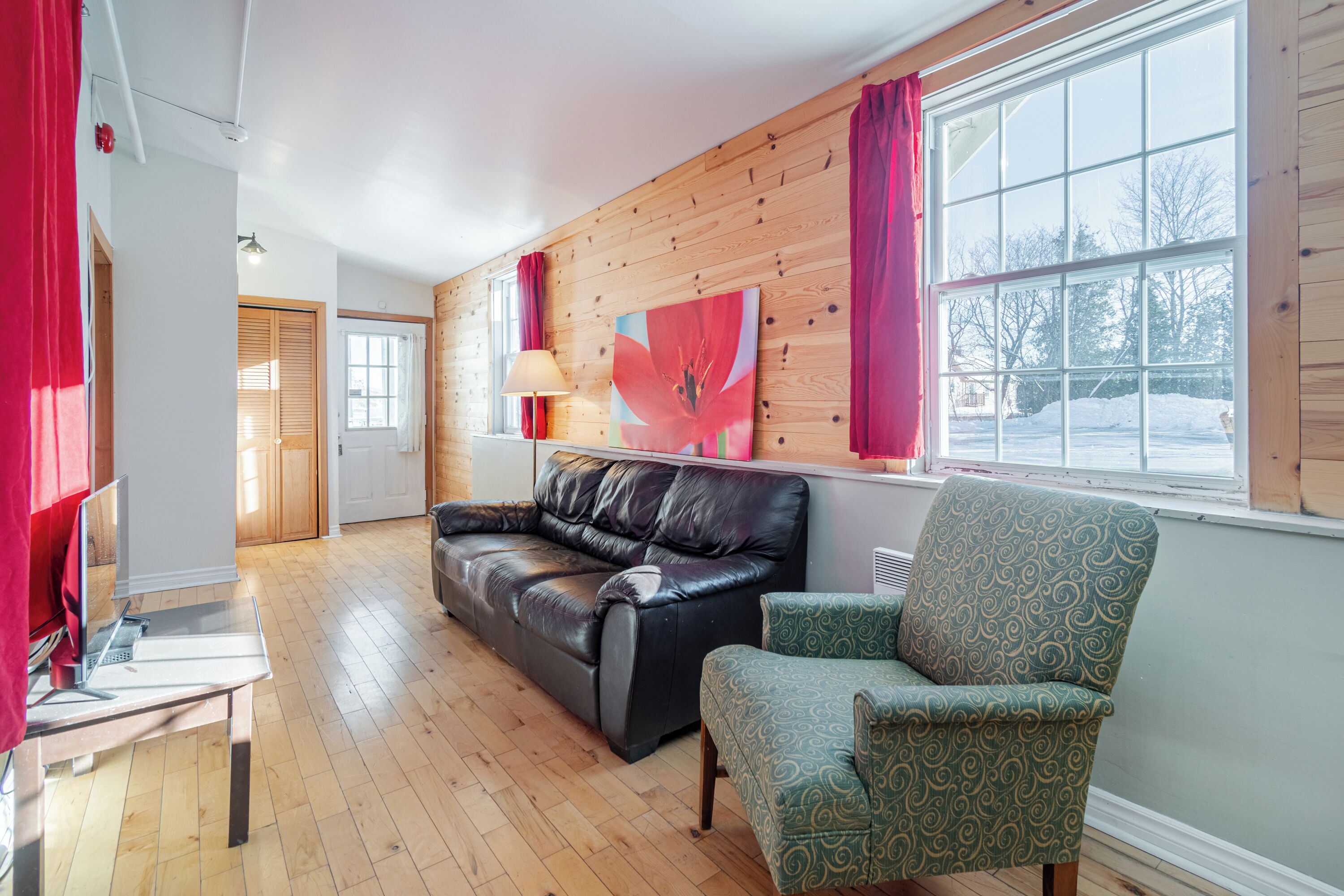 Una habitación con piso de madera, un sofá de cuero negro, un sillón estampado, un cuadro con flores rojas y una ventana que da a un paisaje nevado.