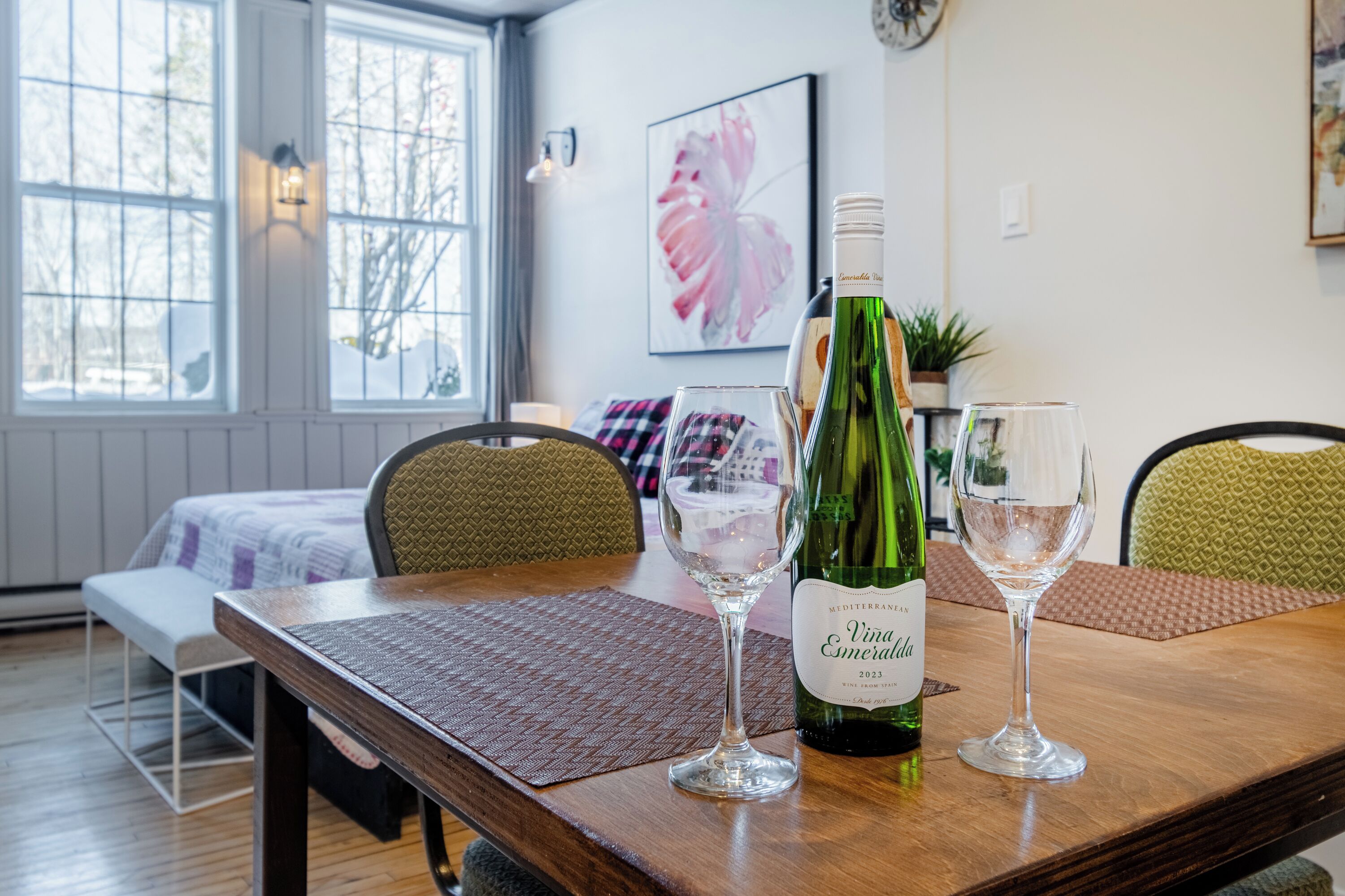 A dining table with a wine bottle and two glasses, a window with a view of trees, and a bed with a checkered blanket.
