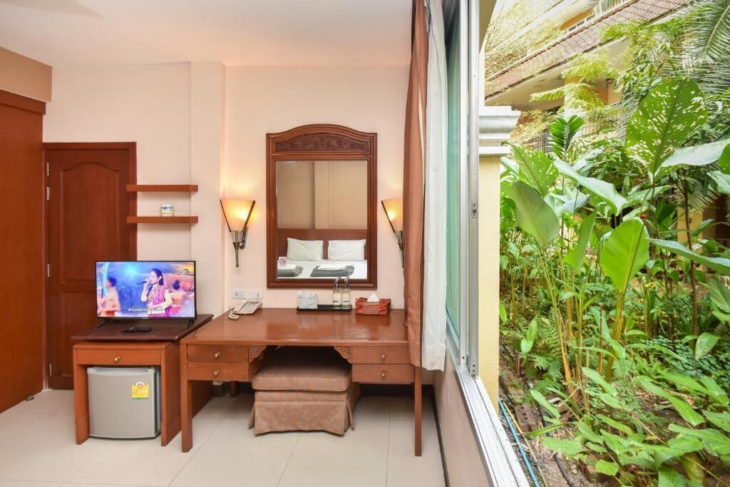 A hotel room with a wooden desk, a television, a mirror, a small refrigerator, and a view of a lush garden.