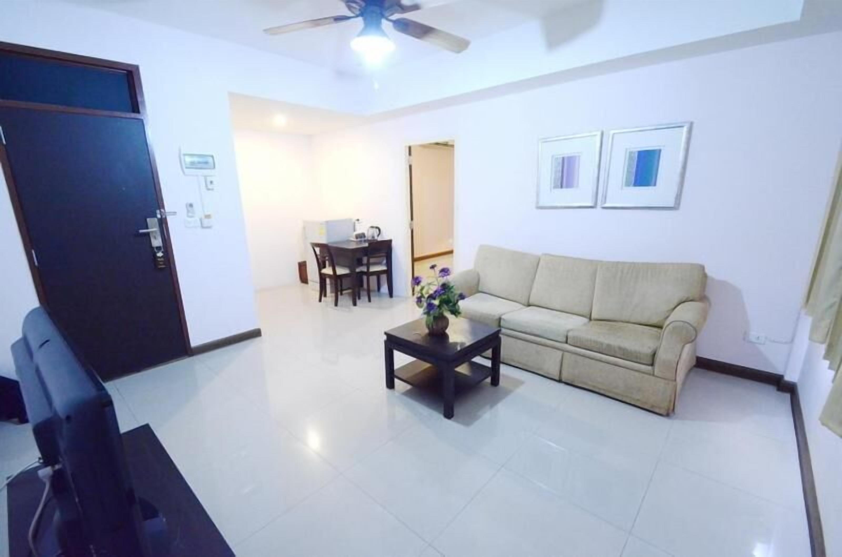 A living room with a sofa, a coffee table, and a TV. There is a door, a dining area, and a ceiling fan.