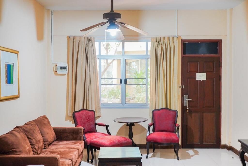 A room with a brown sofa, two red chairs, a small table, a ceiling fan, a window with curtains, and a wooden door.