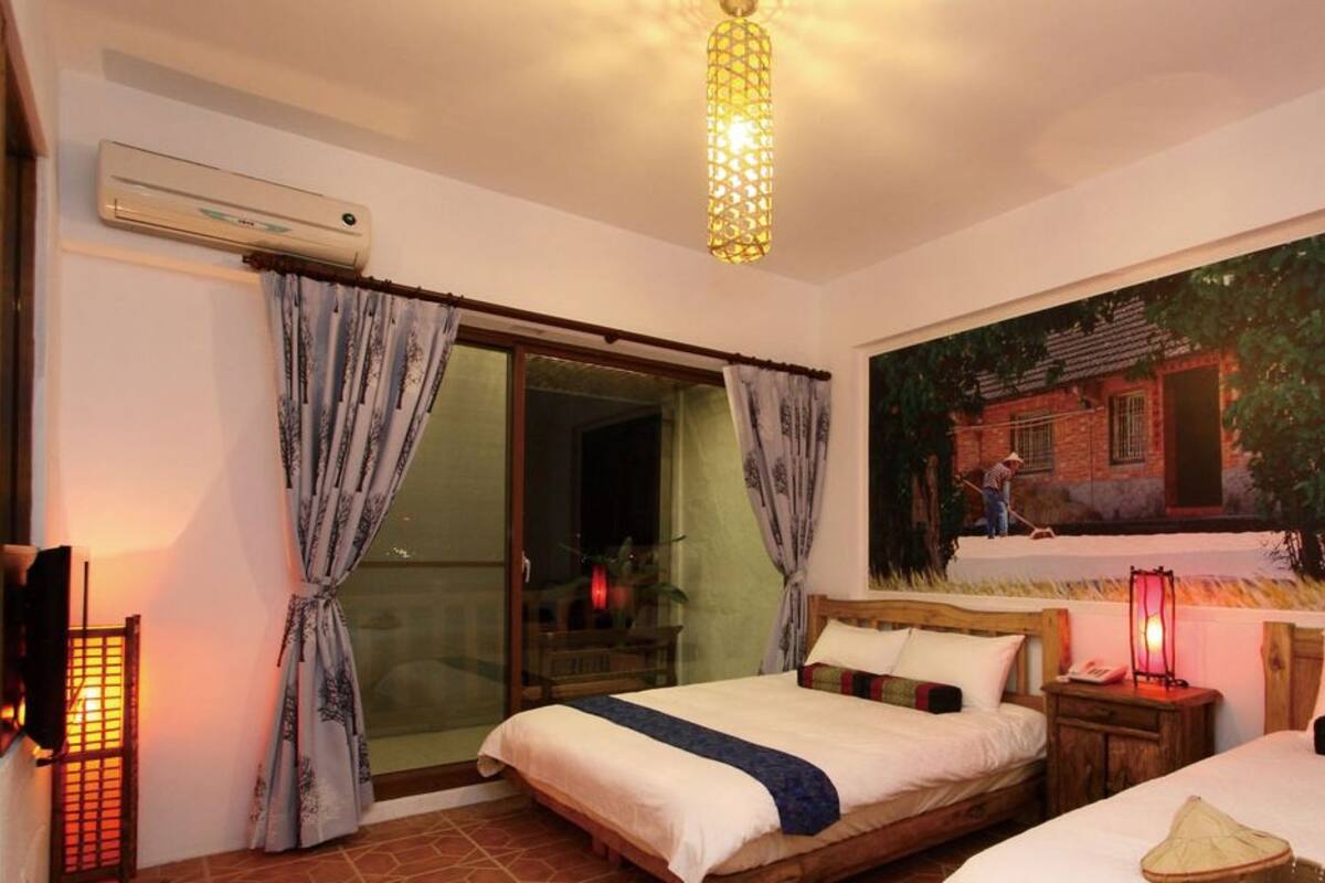 A hotel room with a bed, a nightstand, a wall-mounted air conditioner, a sliding glass door, and a painting of a house.