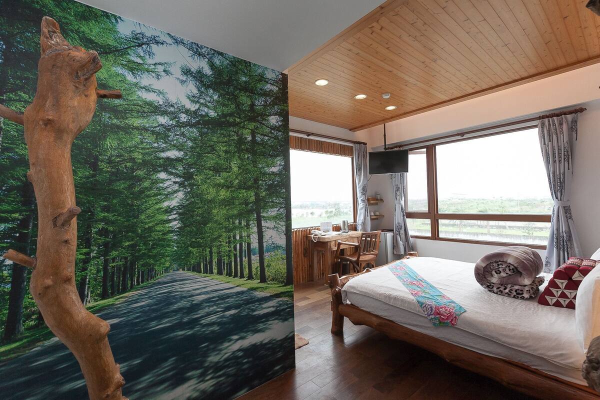 A bedroom with a large window, a bed, a desk, and a view of a forest.