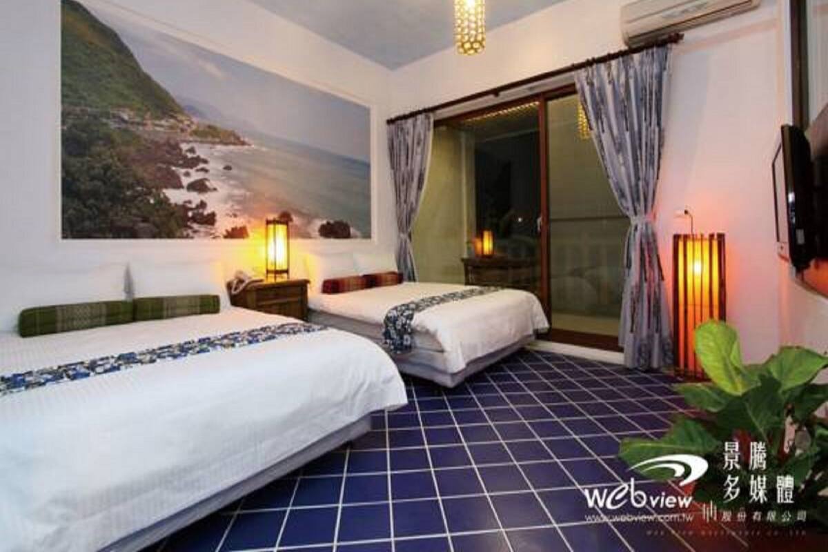 A hotel room with two beds, a large painting of a coastal landscape, a sliding glass door, and a tiled floor.