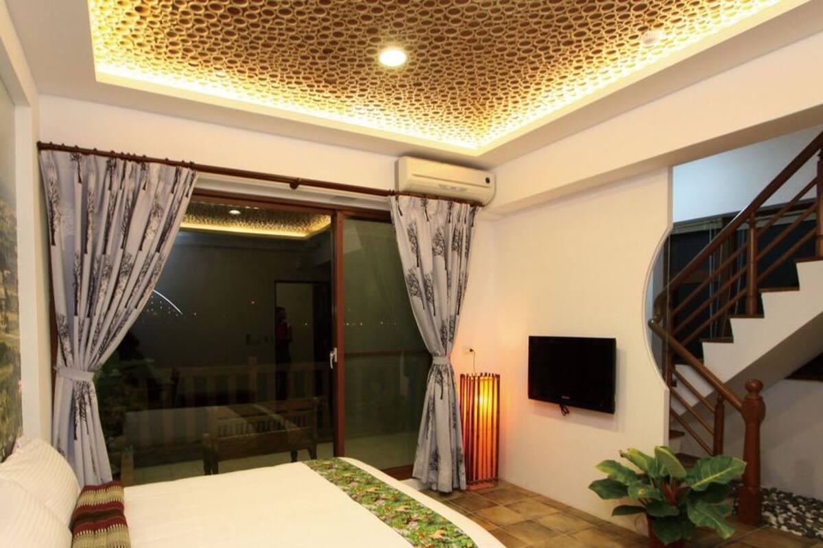 A bedroom with a bed, curtains, a TV, a staircase, and a view of a landscape through a glass door.
