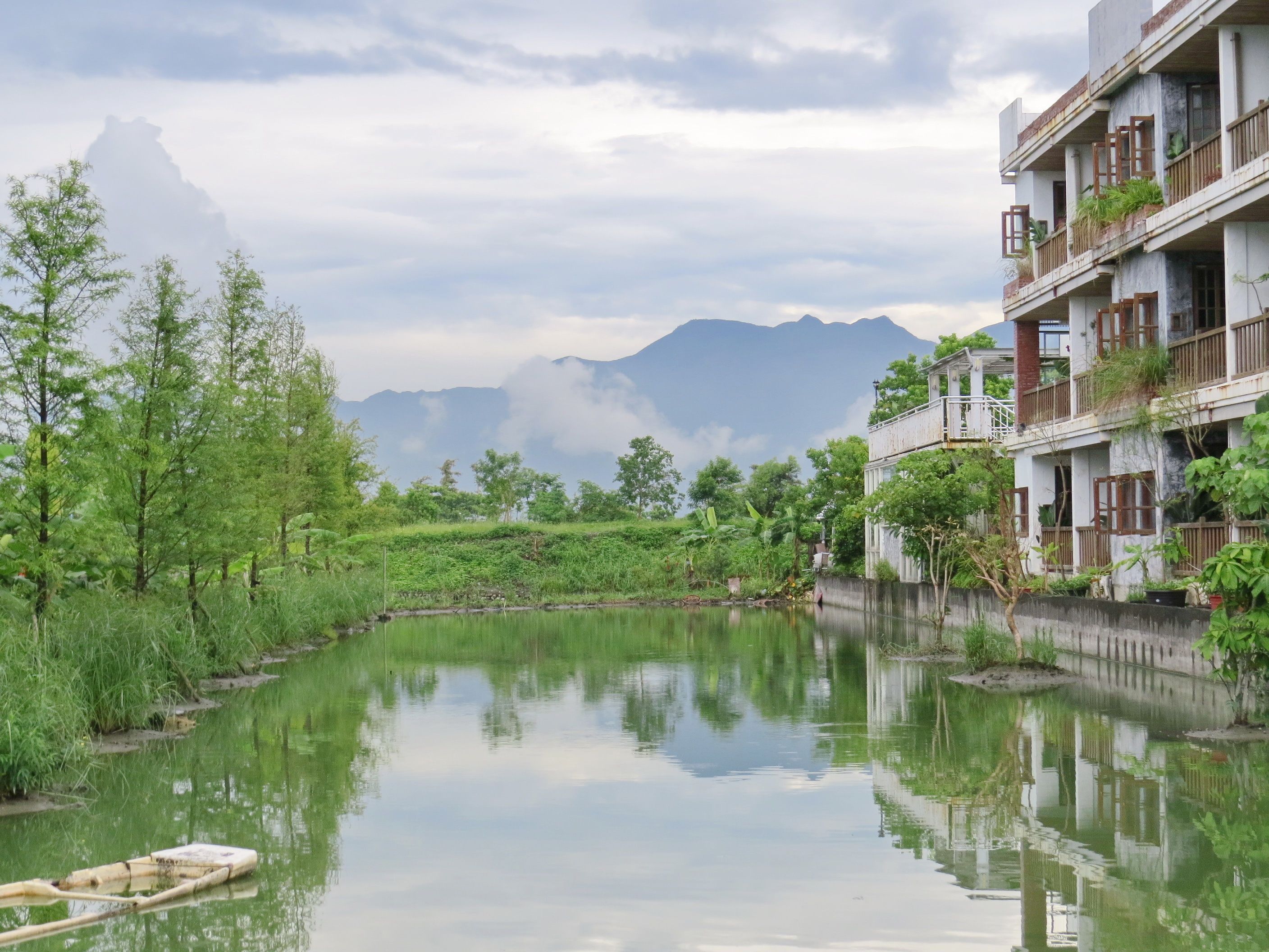A serene riverside view with a building featuring balconies and a reflection of the surrounding greenery and mountains.
