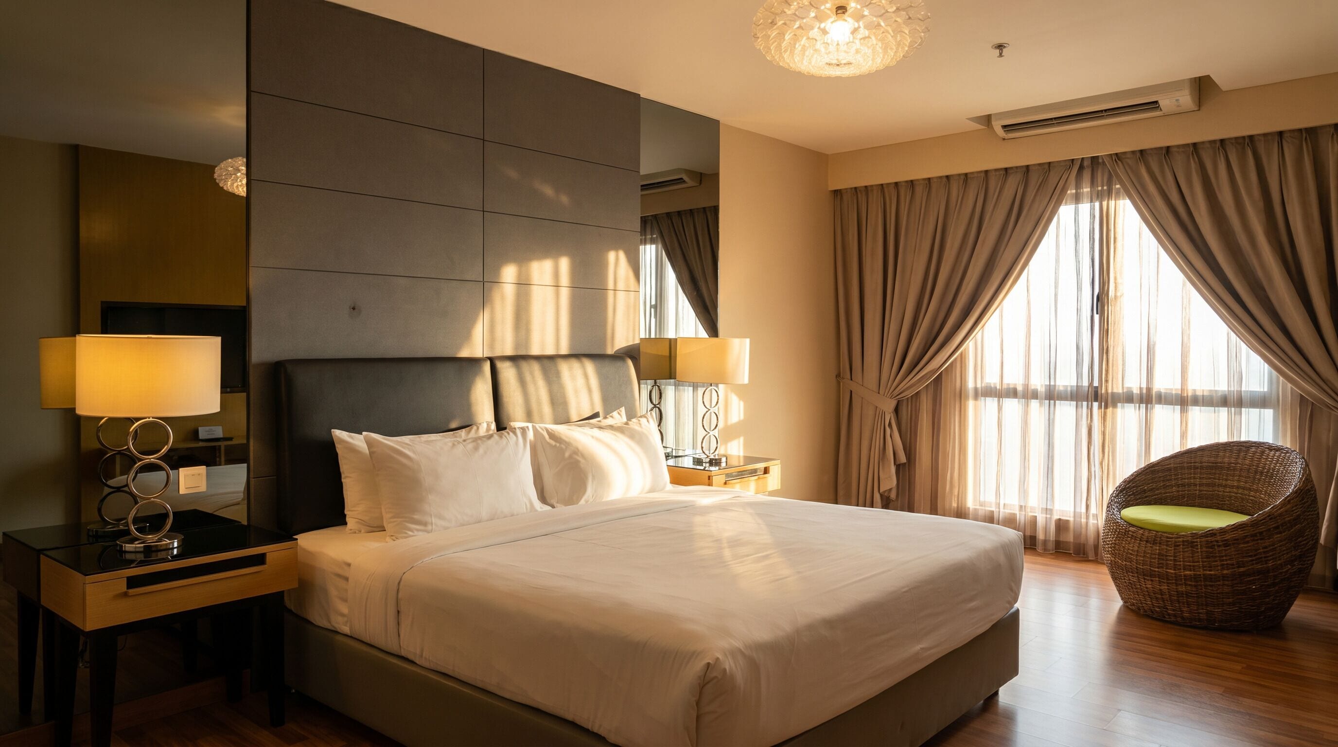 A hotel room with a large bed, two bedside lamps, a wicker chair, and a large window with curtains.