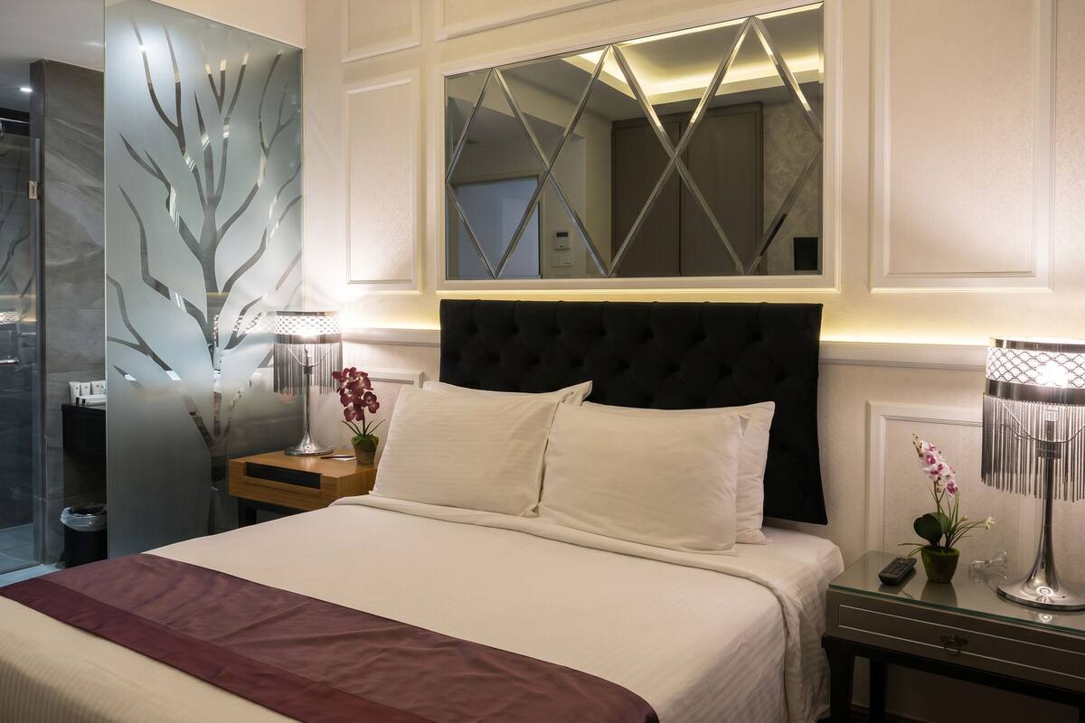 A hotel room with a large bed, a nightstand with a lamp, a mirror, and a window with a geometric design.