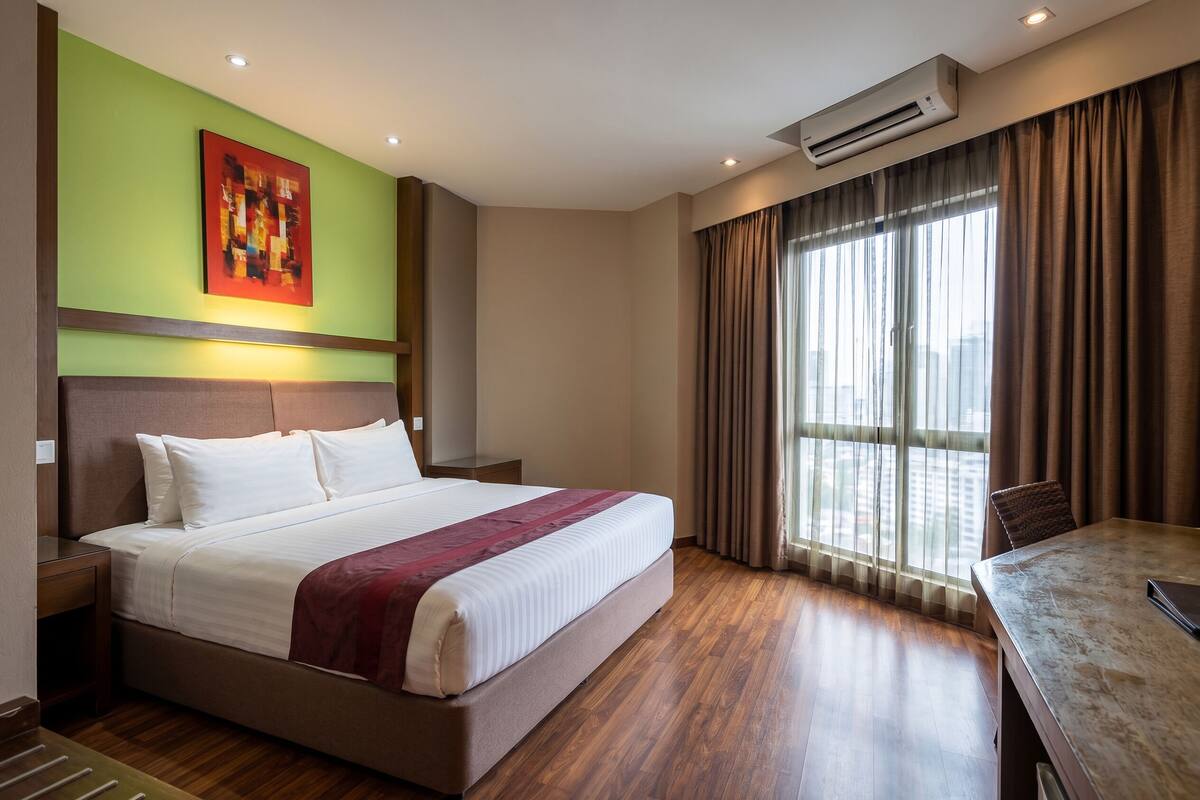 A hotel room with a bed, a dining table, a window with curtains, an air conditioning unit, and a painting on the wall.
