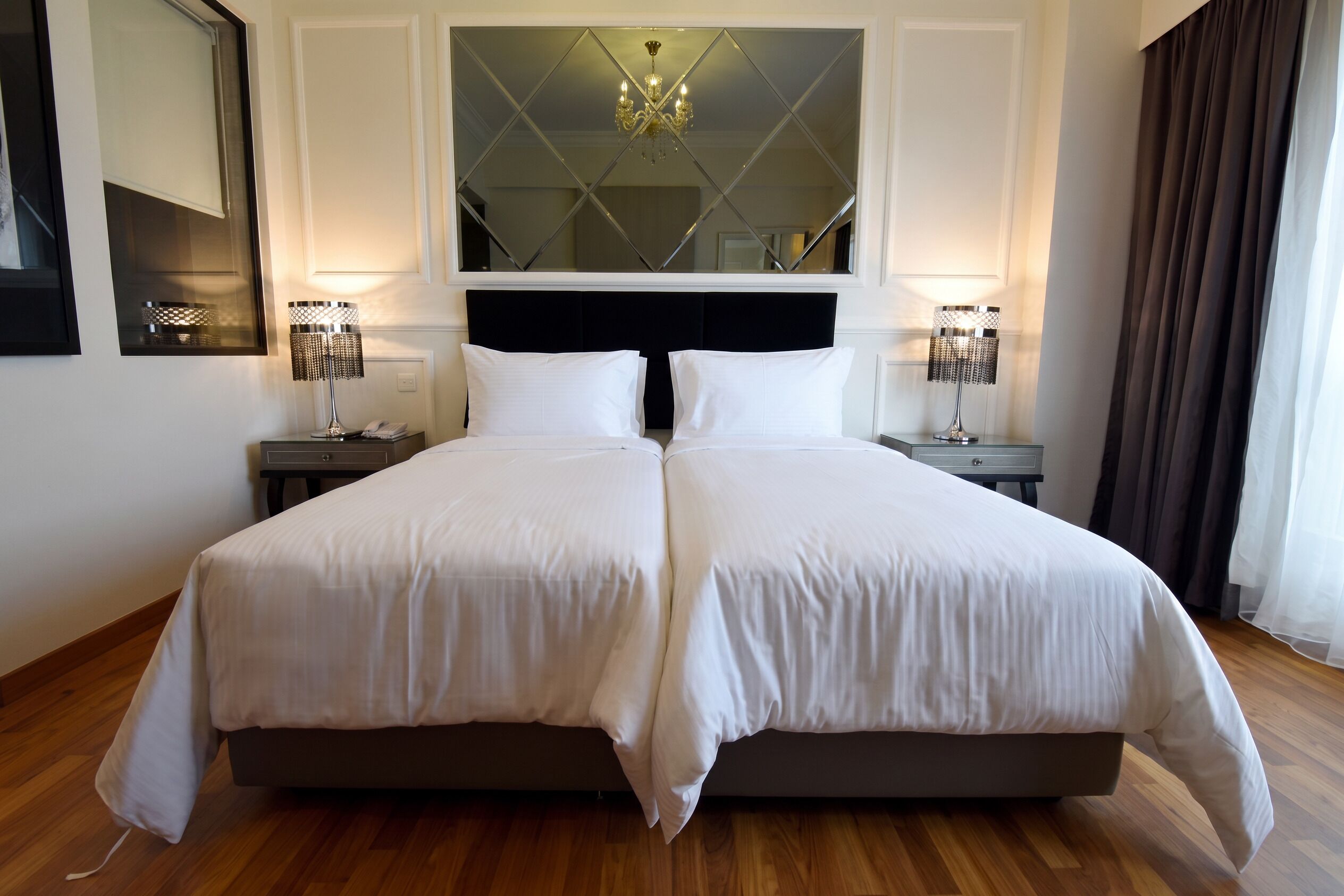 A double bed with white linens, two bedside tables with lamps, a large mirror above the bed, and a chandelier.