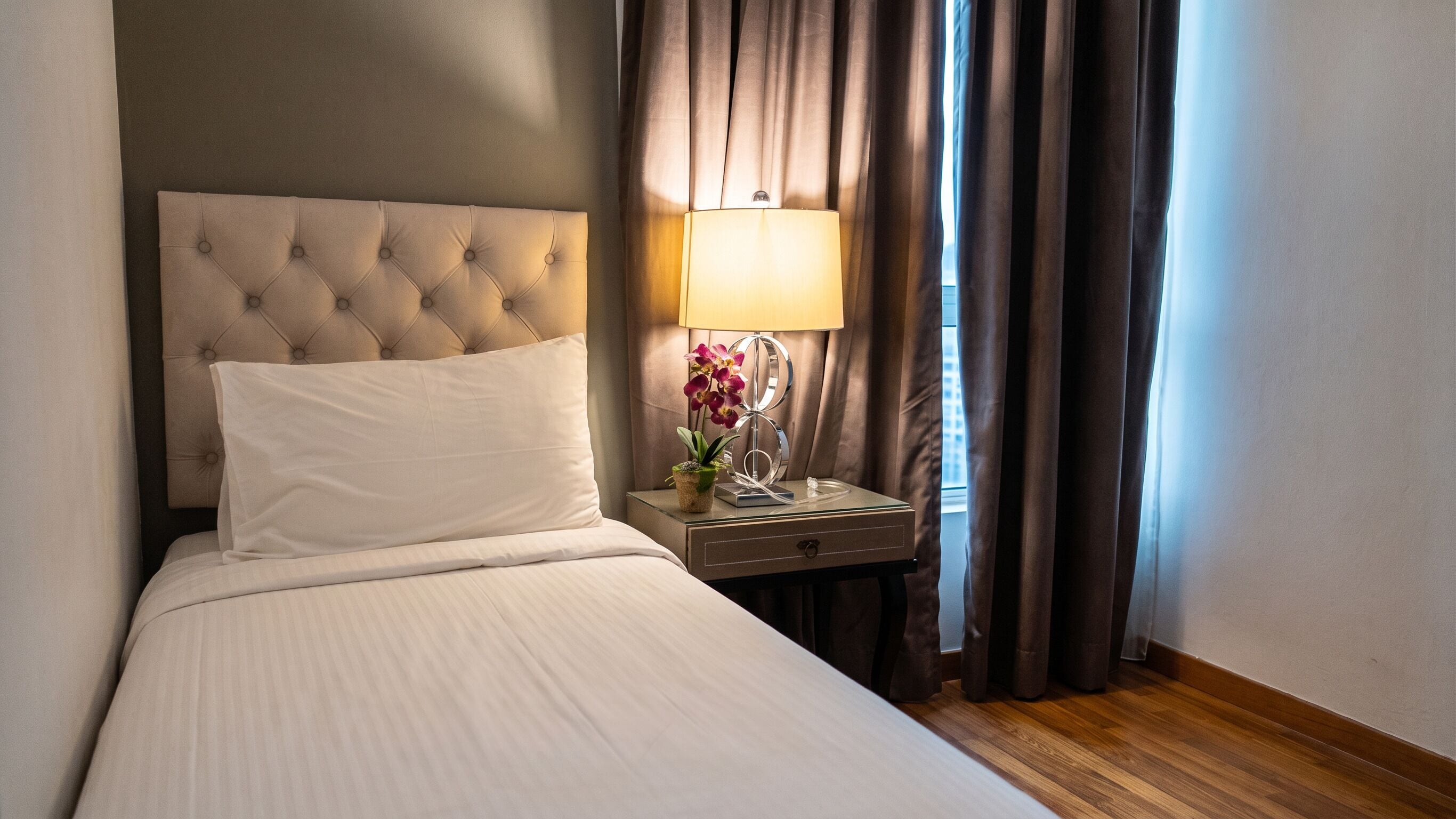 Une chambre d’hôtel avec une tête de lit capitonnée, une table de chevet avec une lampe et des fleurs, et une fenêtre avec des rideaux.