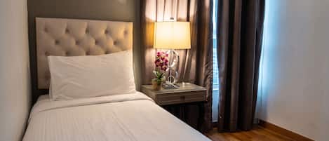 Habitación de hotel con un cabecero capitonado, una mesita de noche con lámpara y flores, y una ventana con cortinas.