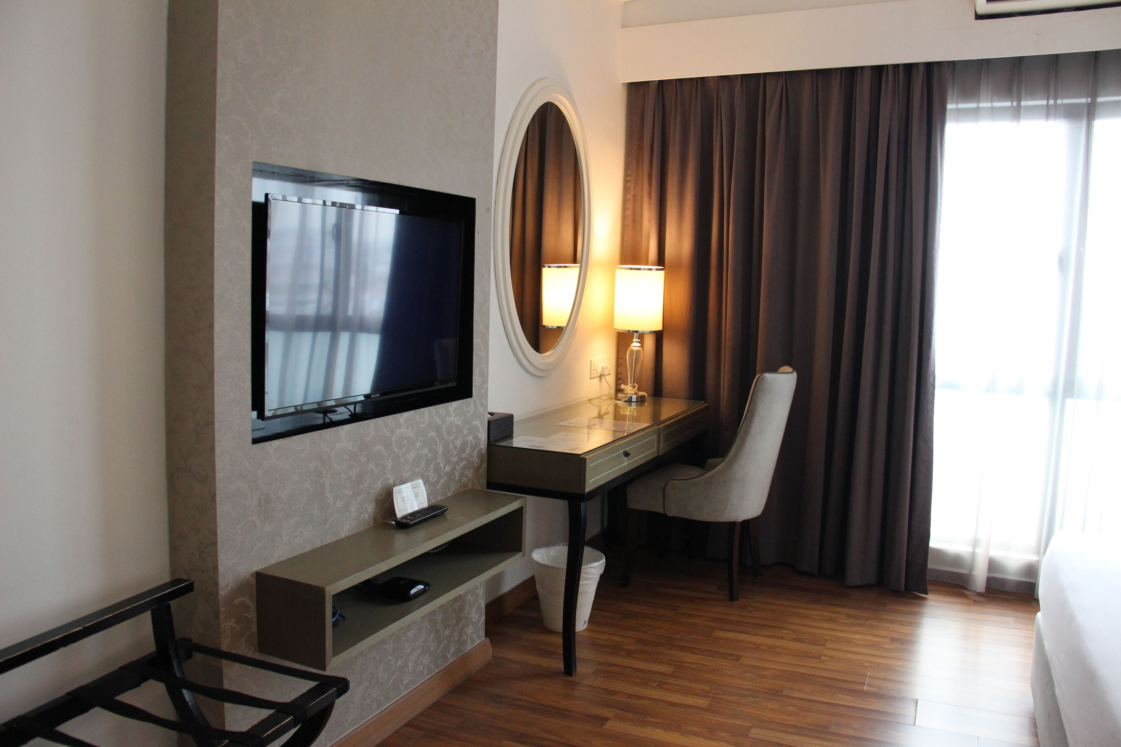 Une chambre d’hôtel équipée d’une télévision, d’un bureau, d’une chaise, d’une lampe et d’une grande fenêtre avec des rideaux.