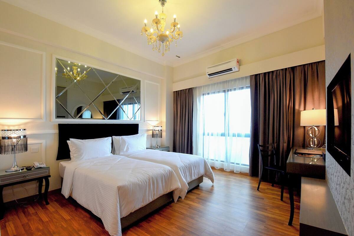 A hotel room with two beds, a chandelier, an air conditioning unit, a desk with a lamp, and a large mirror.