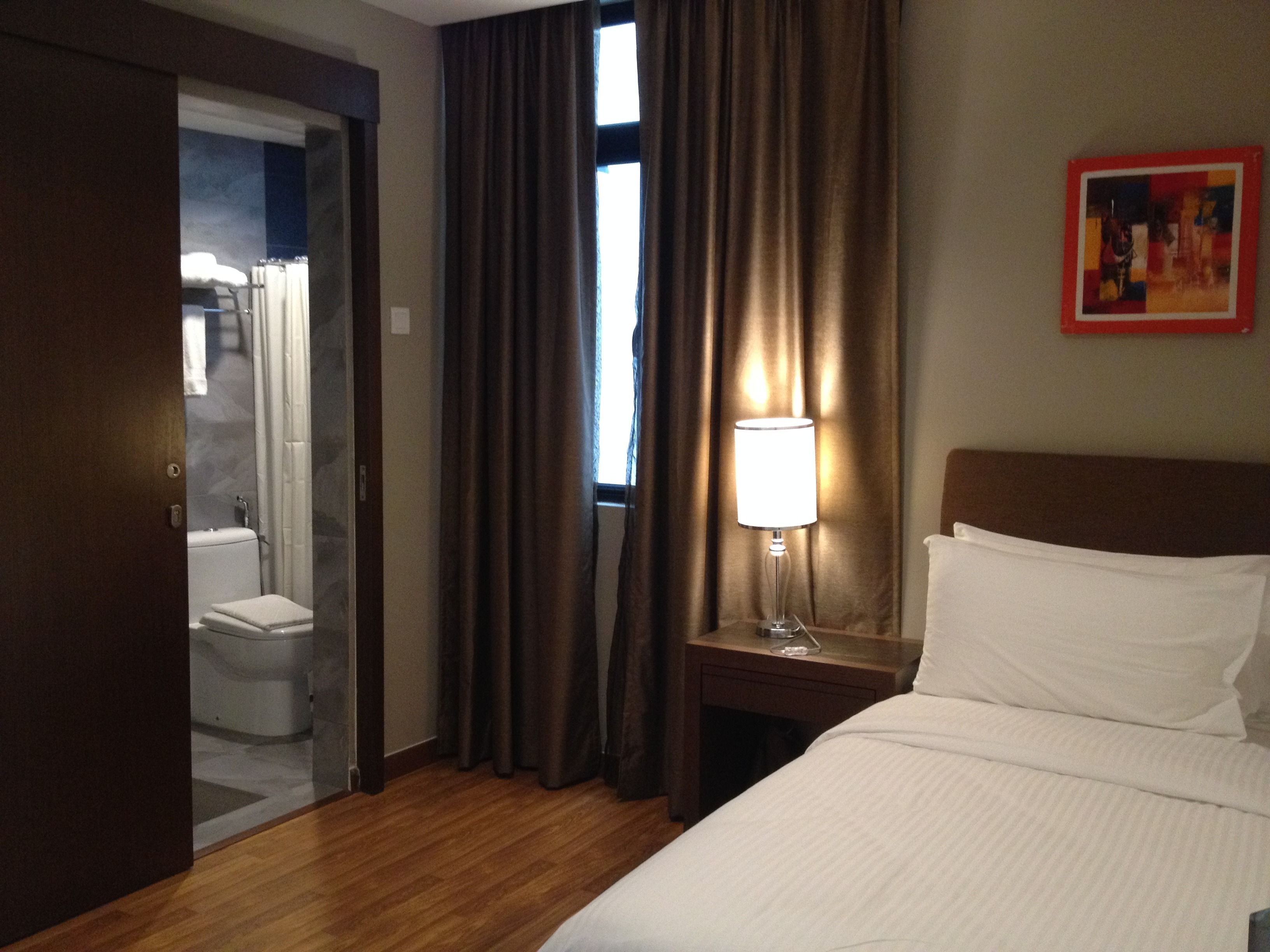 A hotel room with a bed, a nightstand with a lamp, a painting on the wall, and a bathroom visible through an open door.