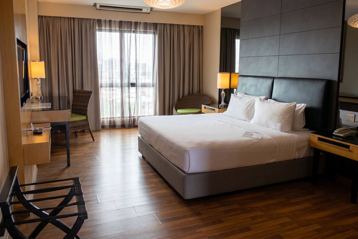 A hotel room with a large bed, a desk with a lamp, a chair, and a view of the outdoors through the window.