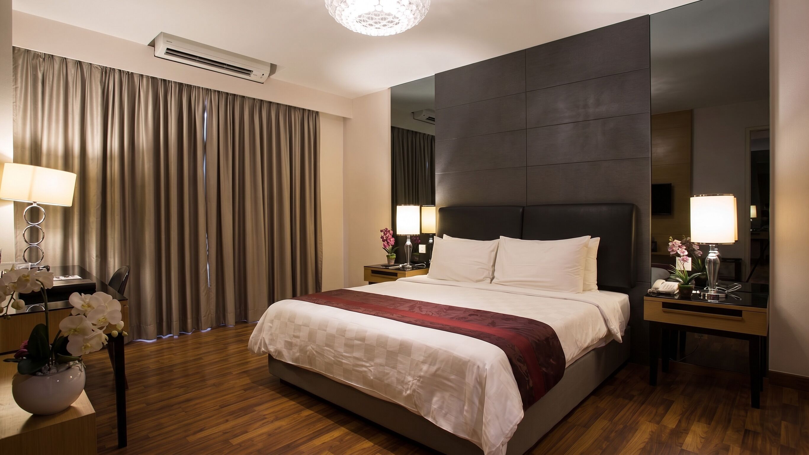 Habitación de hotel con una cama grande, un escritorio con lámpara, una mesita de noche con lámpara y un ventanal con cortinas.
