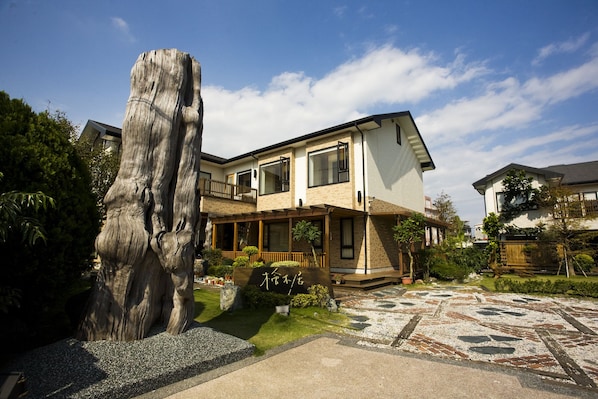 Una casa de dos pisos con un balcón, rodeada por un jardín con un árbol grande y un camino de piedra con diseño.